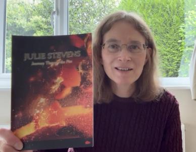 white woman with brown hair wearing glasses in a red jumper holding up a book with an image of flames and the title Julie Stevens Journey Through Fire