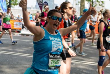 female runner in an MS Trust vest with their hands in the air giving a thumbs up and mouth open