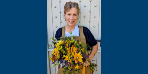 image of a woman wearing a brown apron and a dark blue shirt, holding a bouquet of yellow and purple flowers. She is standing in front of an off white door and has a pair of reading glasses on top of her head.
