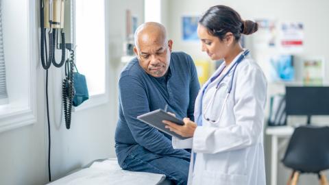 Senior gentleman sits on an exam table during a check-up. His female doctor is holding out a tablet.