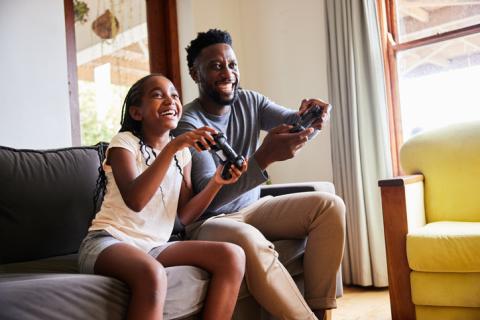 Girl and her father laughing while playing video games together on a sofa in their living room