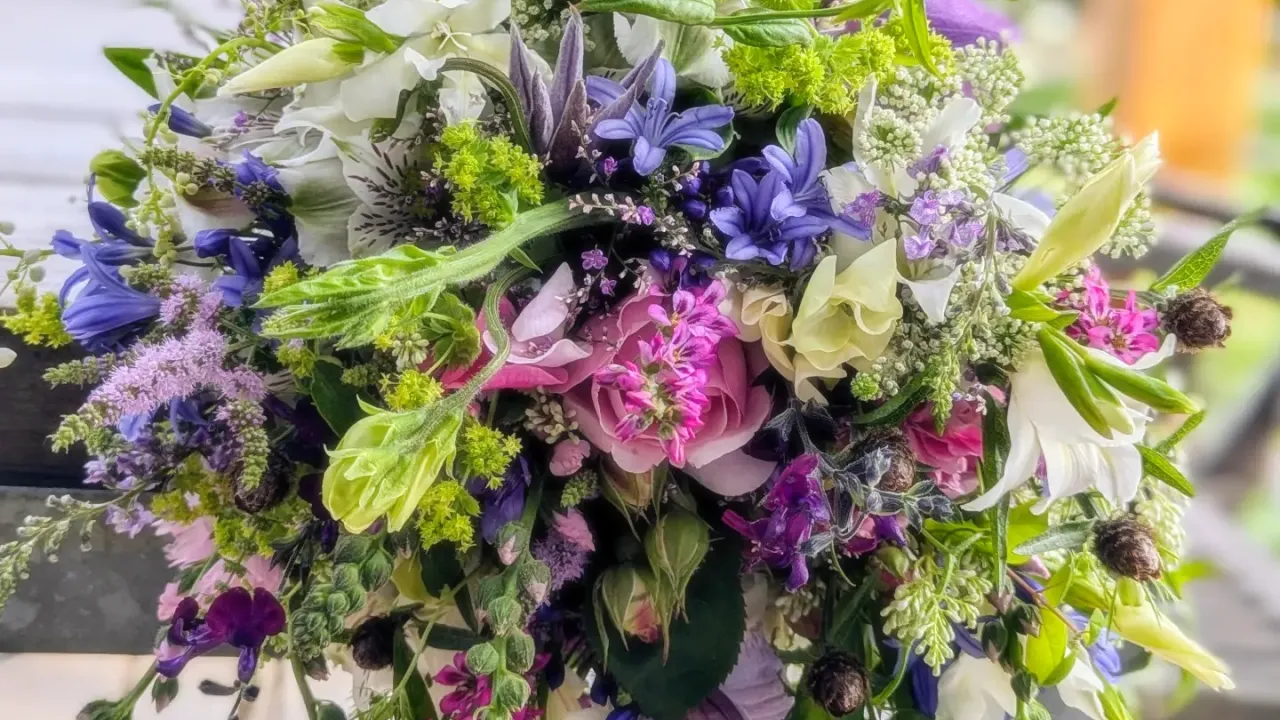 A vibrant, cascading bouquet featuring a mix of pastel and bold flowers, including pink roses, purple clematis, white lilies, and delicate green foliage, arranged in a natural, flowing style.