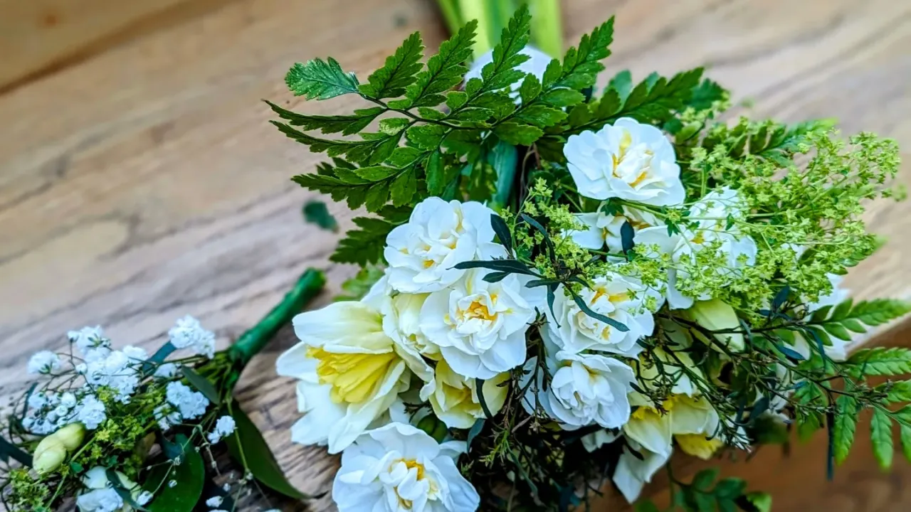 Bouquet of white flows and white daffodils and green foliage fern plants wrapped up and lying on a step facing towards trhe camera. Beside is a smaller bouquet, about a quarter of the size, of baby's breath flowers and leaves