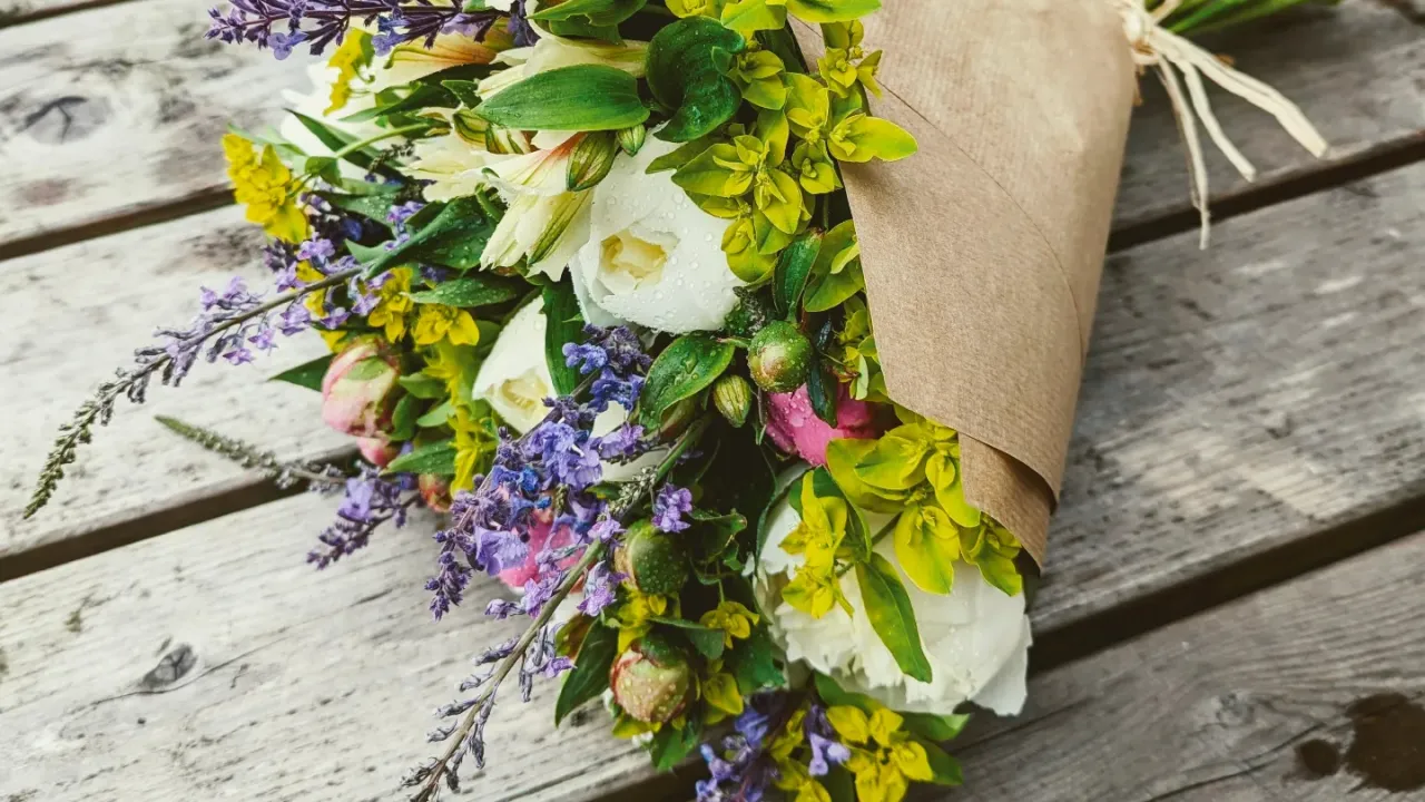 Large bouquet of flowers lying on its size on a wooden flooring. The flowers are white, purple, pink and there is a lot of green foliage too. It is wrapped in brown paper and tied with a strong. It sits facing the bottom left corer of the photo. 