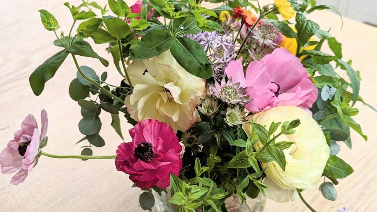 Glass vase sat on a wooden table containing a small bunch of flowers which are pink, white and yellow surrounded by green foliage.